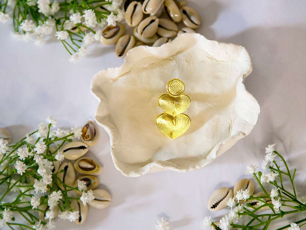 Bague dorée en forme de coeur posée sur un plateau blanc avec un fond de décor de cauris et fleure blanche