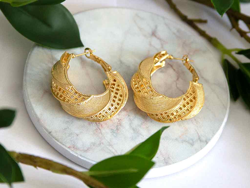 Grosse boucles d'oreilles dorées en forme de boucle posées sur un plateau de marbre avec des feuilles verts en arrière plans