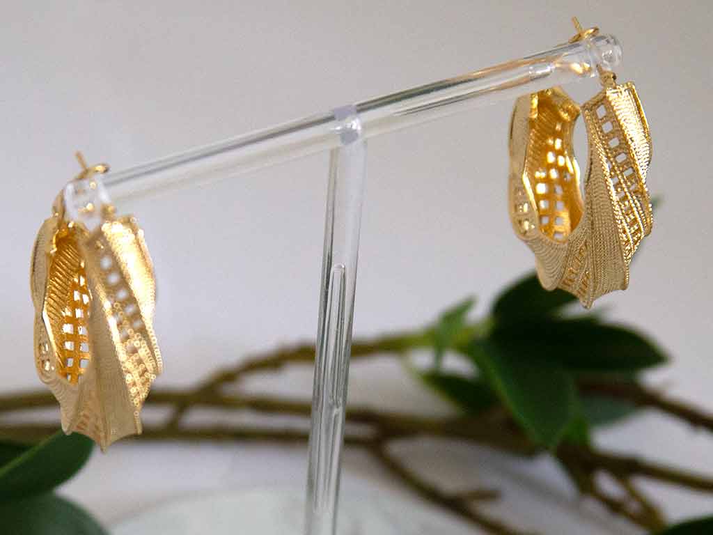 Grosse boucles d'oreilles dorées en forme de boucle posées sur un présentoir  avec des feuilles verts en arrière plans