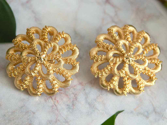Grosses boucles d'oreilles dorées en forme de fleur posées sur un plateau en marbre avec des feuilles vertes en arrière plan