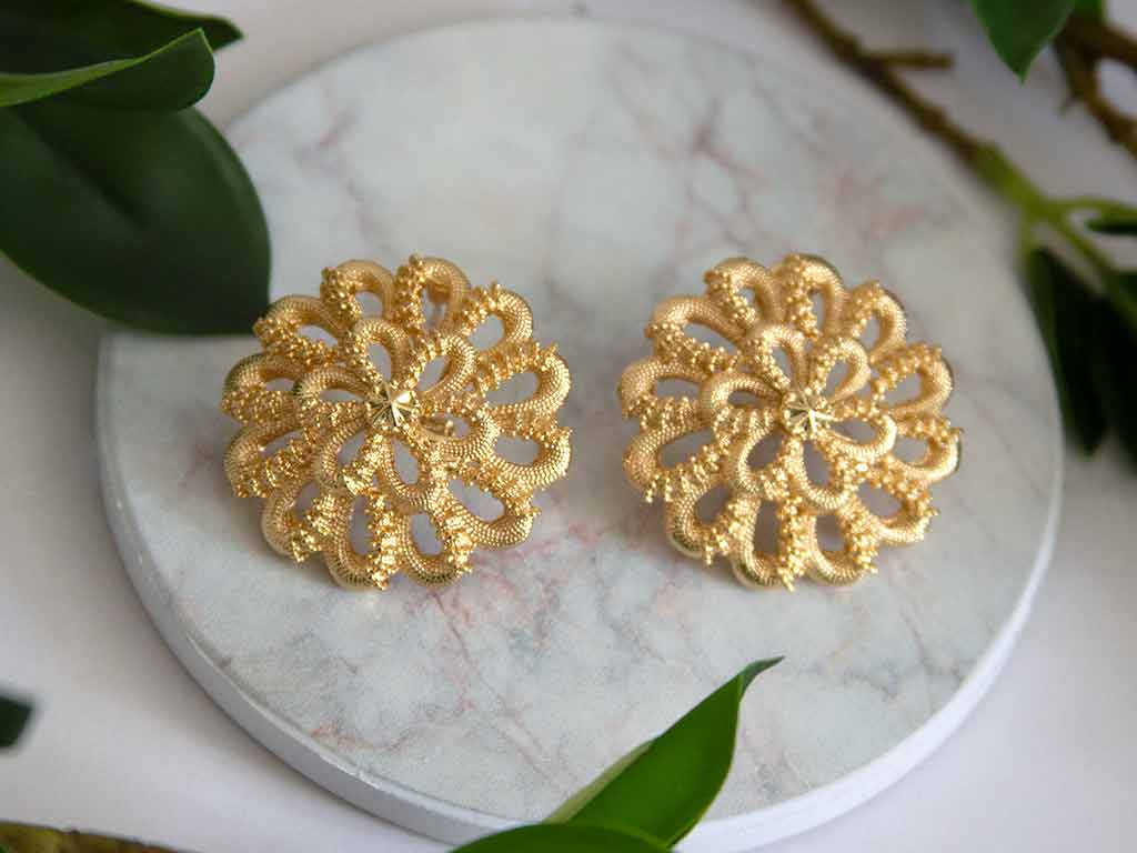Grosses boucles d'oreilles dorées en forme de fleur posées sur un plateau en marbre avec des feuilles vertes en arrière plan