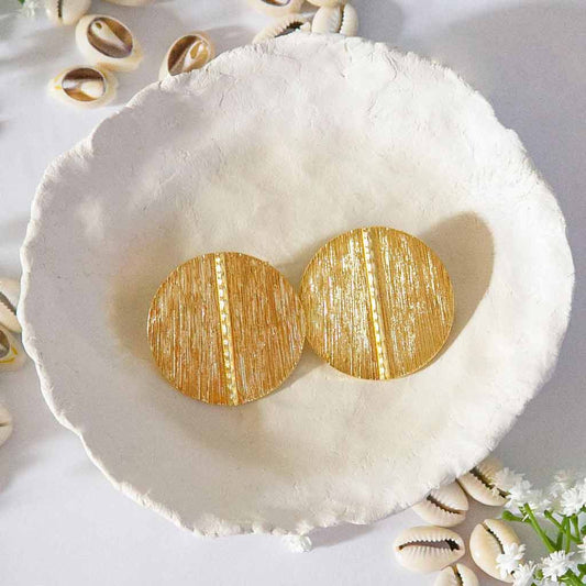Boucles d'oreilles ethniques, rondes dorées, posées sur un plateau avec des coquillages cauris et des fleures blanches en décor