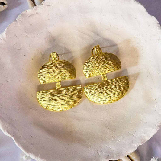 Boucle d'oreille de style ethnique dorées en forme de deux demi-cercle, posées sur un plateau avec un décor de coquillage cauris et des fleures blanches