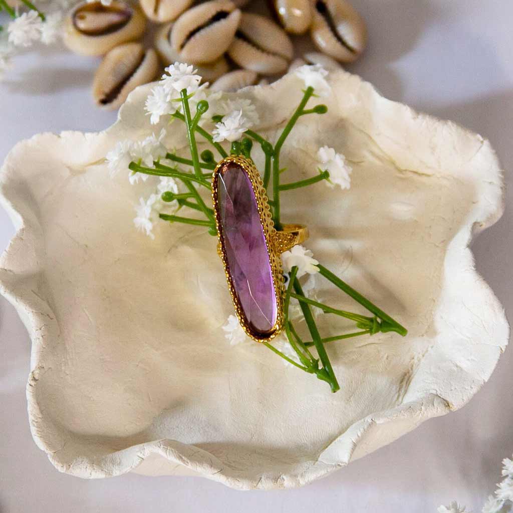 Une Bague avec un cristal violet posée sur une coupelle avec des fleurs blanches et coquillages cauris en décor