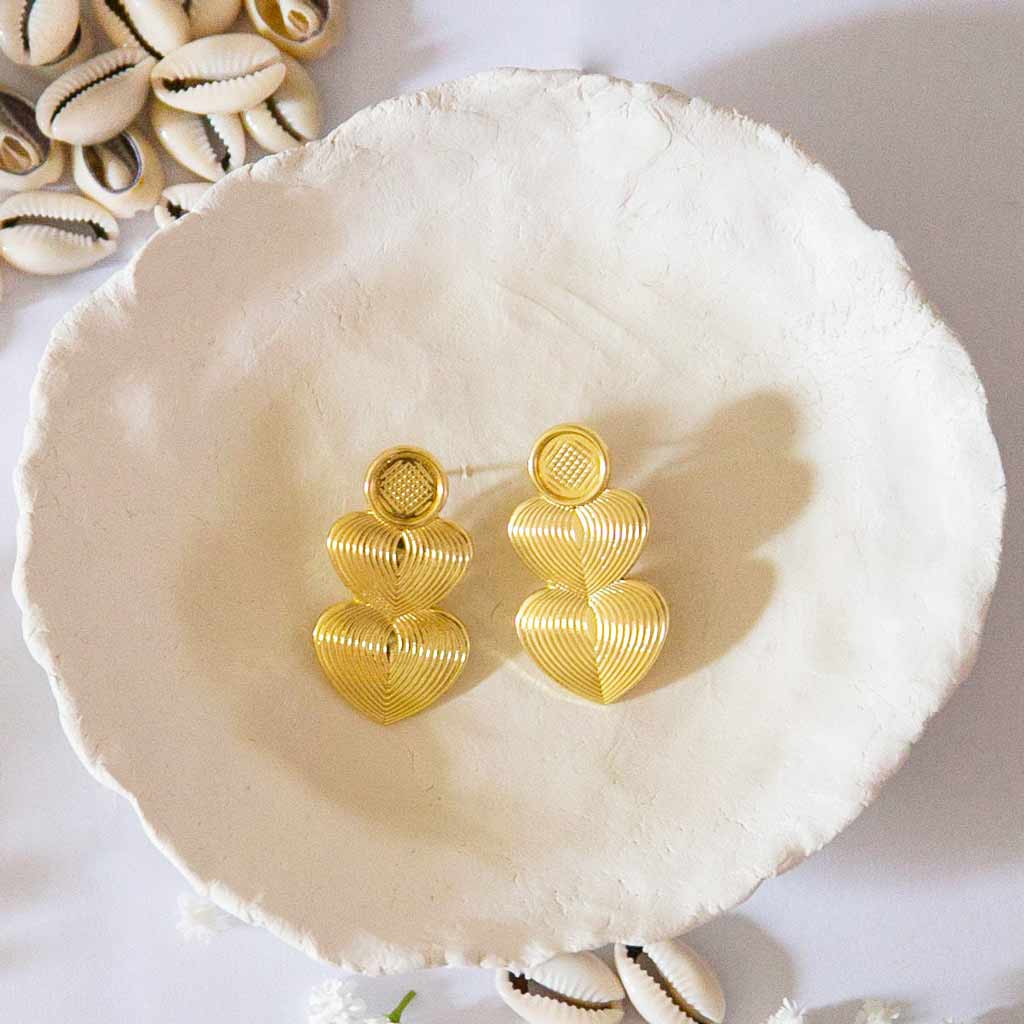 Boucles d'oreilles dorées, longues, en forme de coeurs, posées sur un plateau avec un décor de coquillages cauris et des fleures blanches
