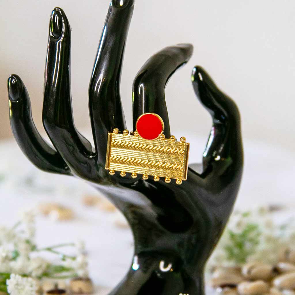 Main de mannequin portant, à l'index, une bague rectangulaire, dorée et rouge, avec un décor de coquillage cauris et des fleures blanches.