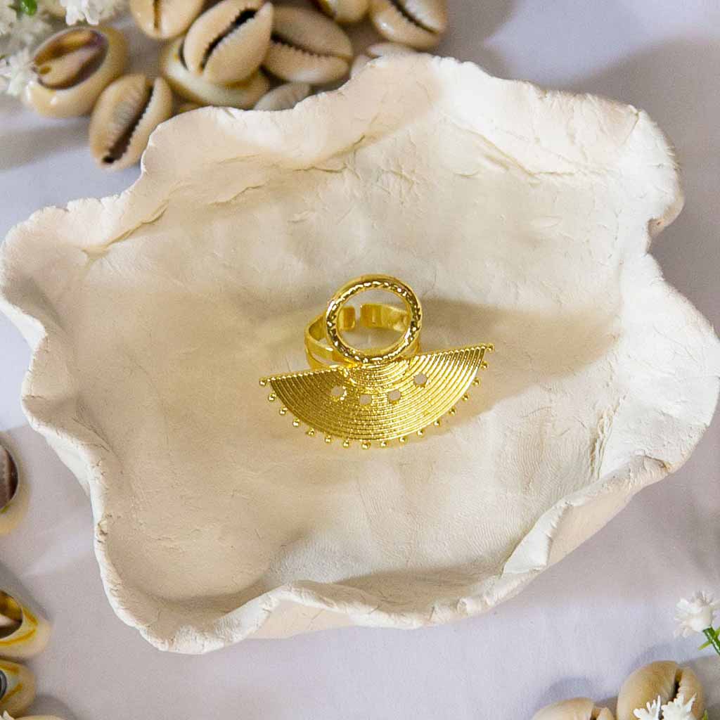 Bague au style ethnique, dorée en forme de demi rond, posée sur un plateau avec un décor de coquillage cauris et des fleurs blanches