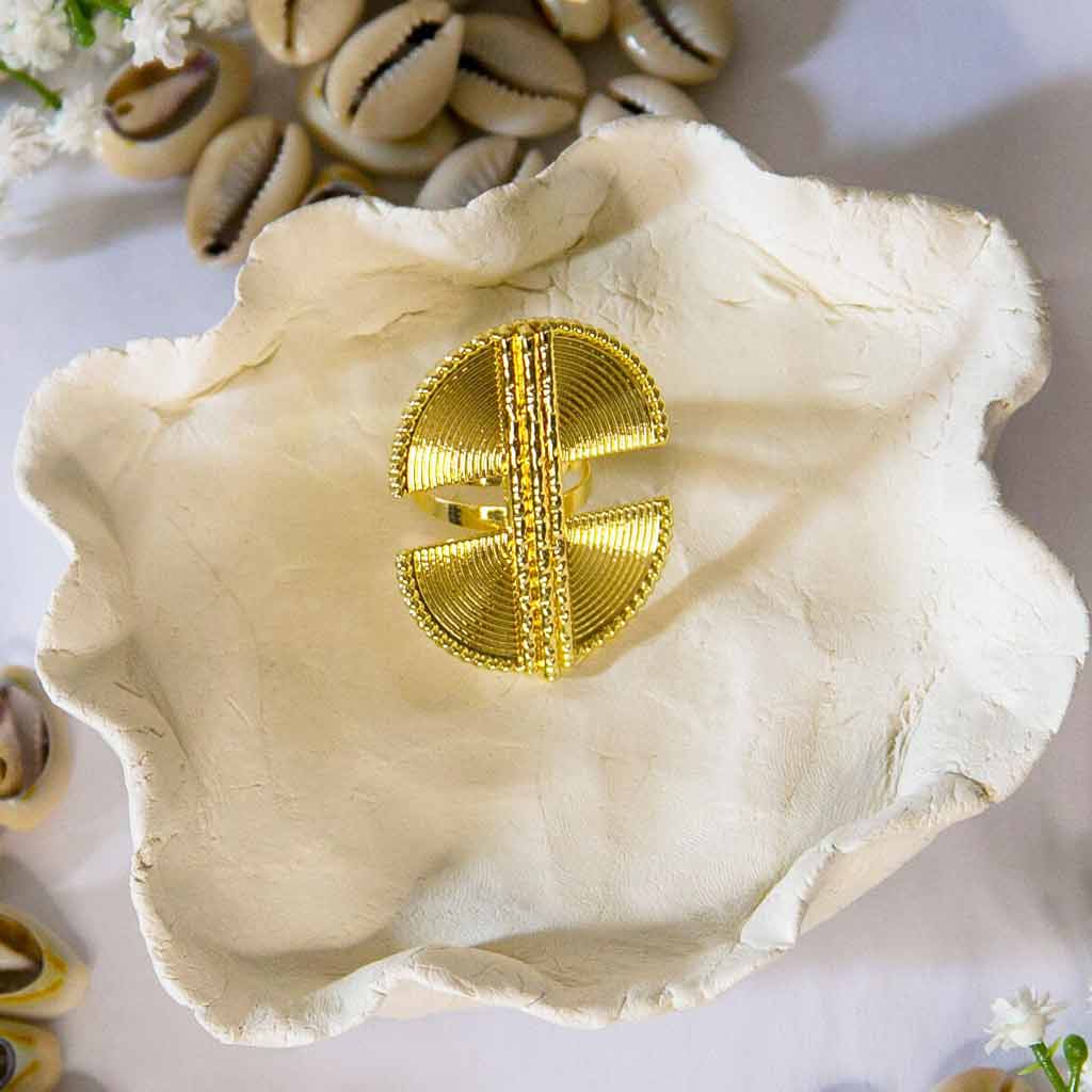 Bague dorée, de style ethnique, posée sur un plateau avec un décor de coquillage cauris et de fleurs blanches