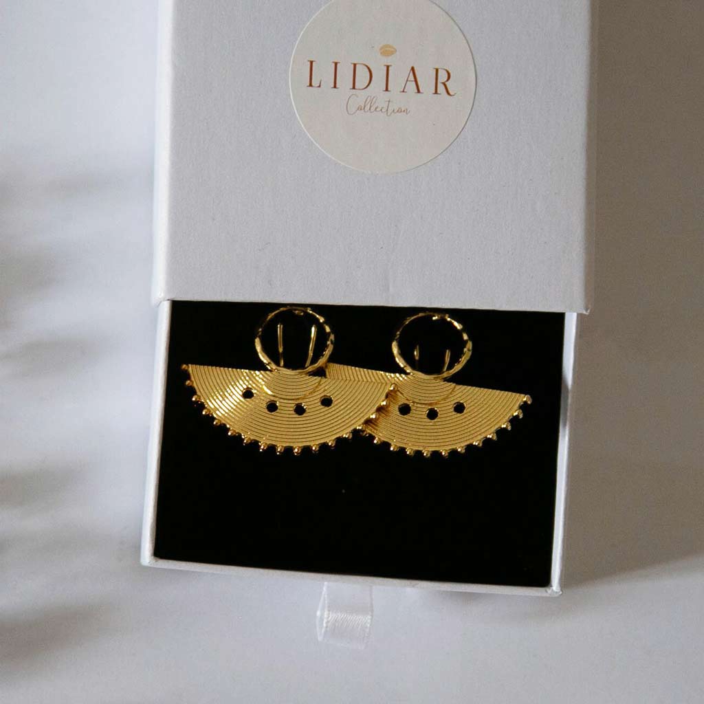 Boucles d'oreilles au style ethnique, dorées en forme de demi rond, posées dans un écrin à bijoux blanc avec le logo lidiar collection
