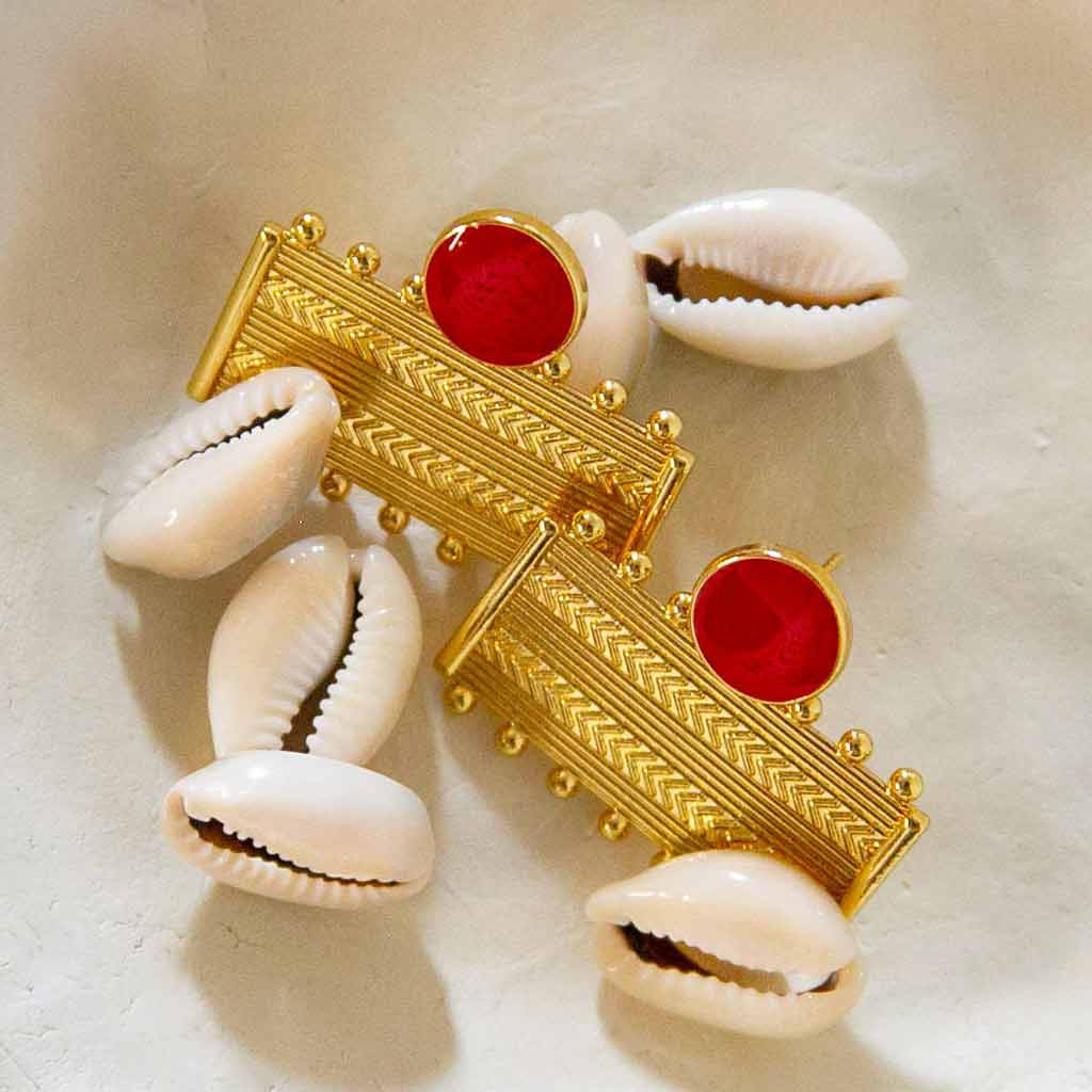Boucles d'oreilles dorées et rouge, rectangulaires, avec des formes géométriques, posées un plateau avec des coquillages cauris