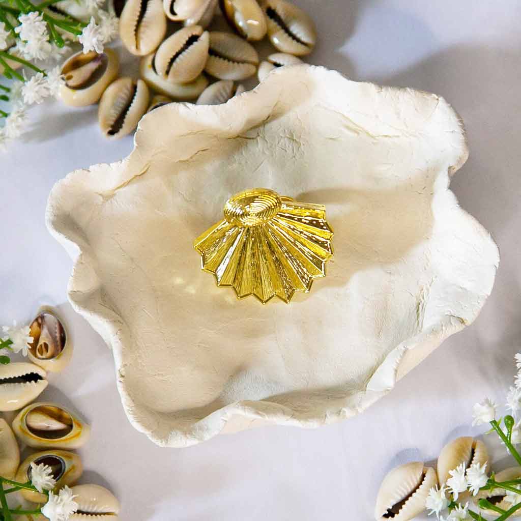 Bague de style ethnique, dorée, posée sur un plateau avec un décor de coquillage cauris et de fleures blanches