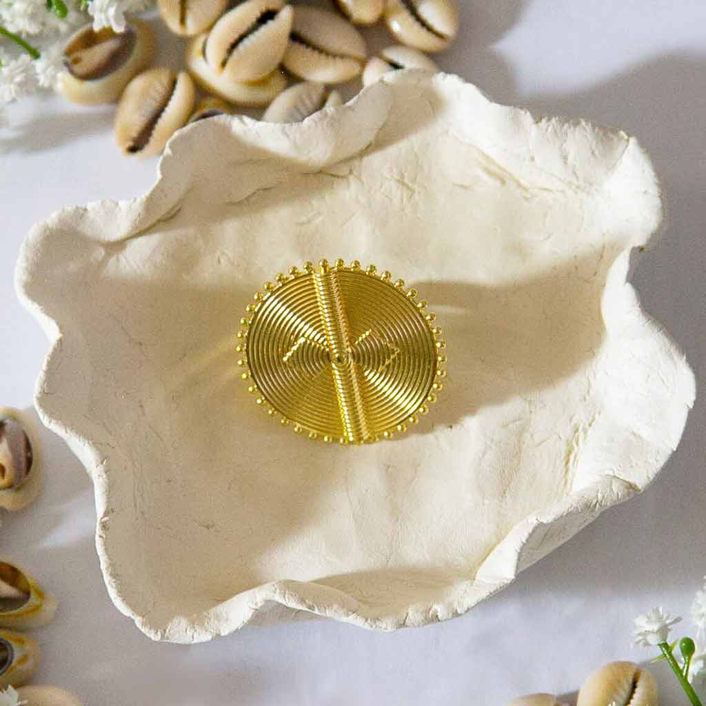 Bague ronde , avec des motifs ethniques, dorée, posée sur un plateau avec des coquillages cauris et des fleures blanches en décors