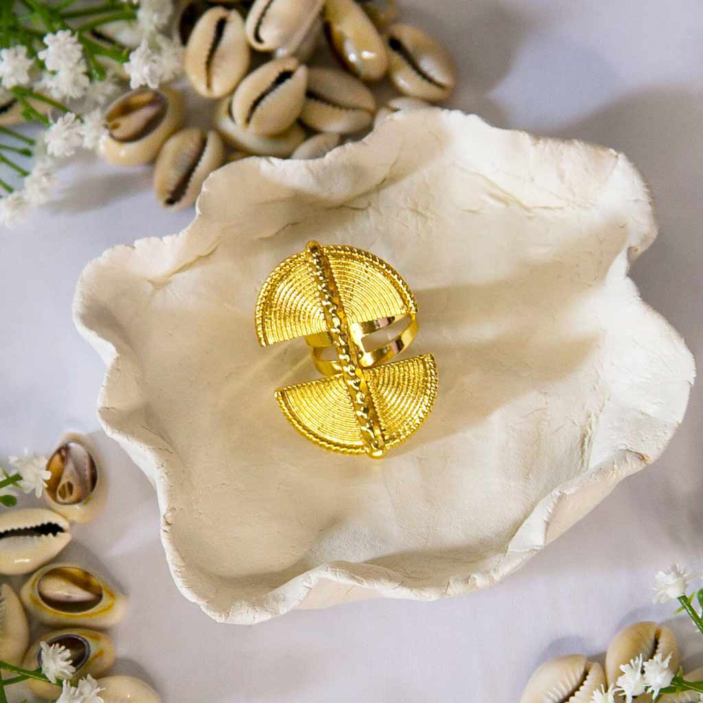Une bague dorée , de style ethnique, posée sur un plateau avec un décor de coquillages cauris et de fleurs blanches