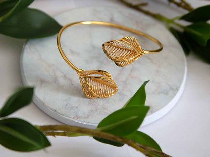 bracelet doré en forme de feuille posé sur un plateau de marbre avec des plantes vertes en décor