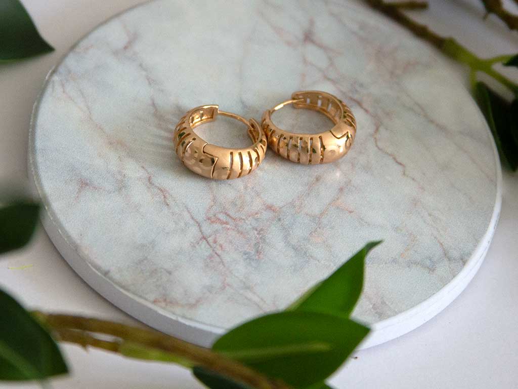 petite boucles d'oreilles dorées posée sur un plateau de marbre avec des feuilles vertes en arrière plan