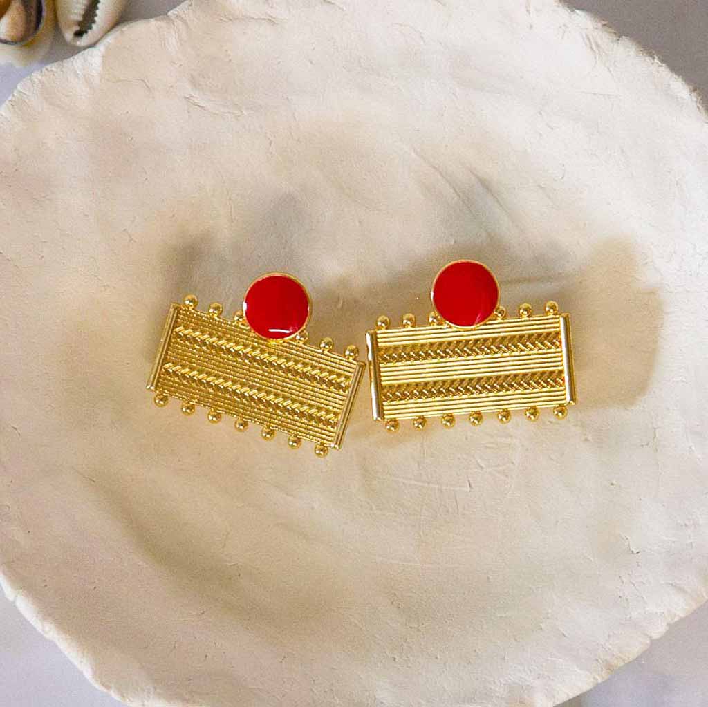 Boucles d'oreilles dorées et rouge, rectangulaires, avec des formes géométriques, posées un plateau avec des coquillages cauris et des fleures blanches en décor
