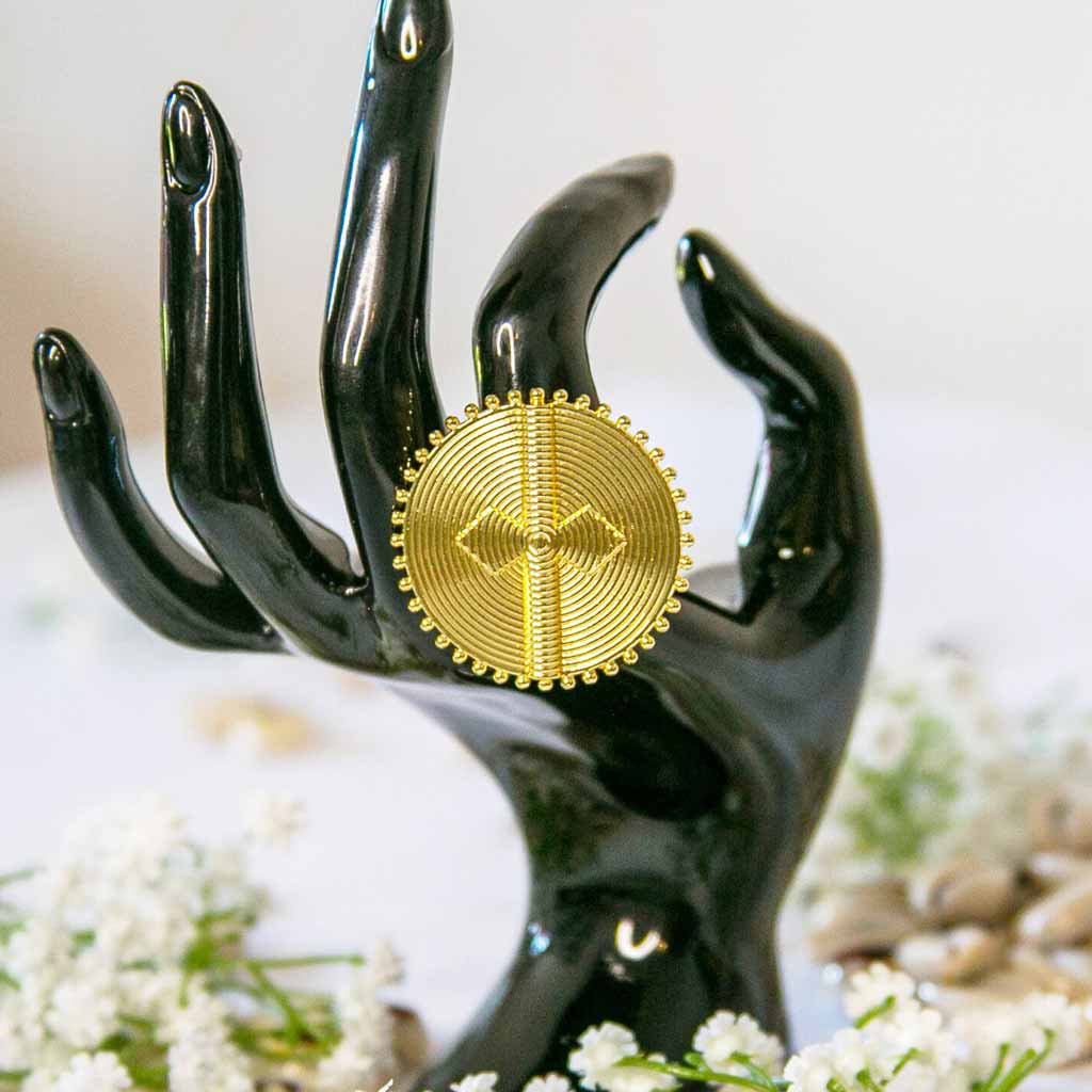 Main de mannequin portant une bague, à l'index, de style ethnique , ronde et dorée, avec un décor de coquillage cauris et des fleures blanches