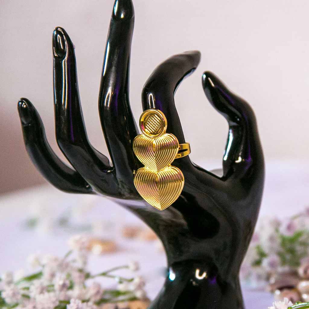 Main de mannequin portant à l'index une bague dorée longue avec des coeurs, avec un décor en fond de coquillage cauris et de fleurs blanches