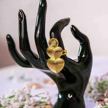Main de mannequin portant à l'index une bague dorée longue avec des coeurs, avec un décor en fond de coquillage cauris et de fleurs blanches