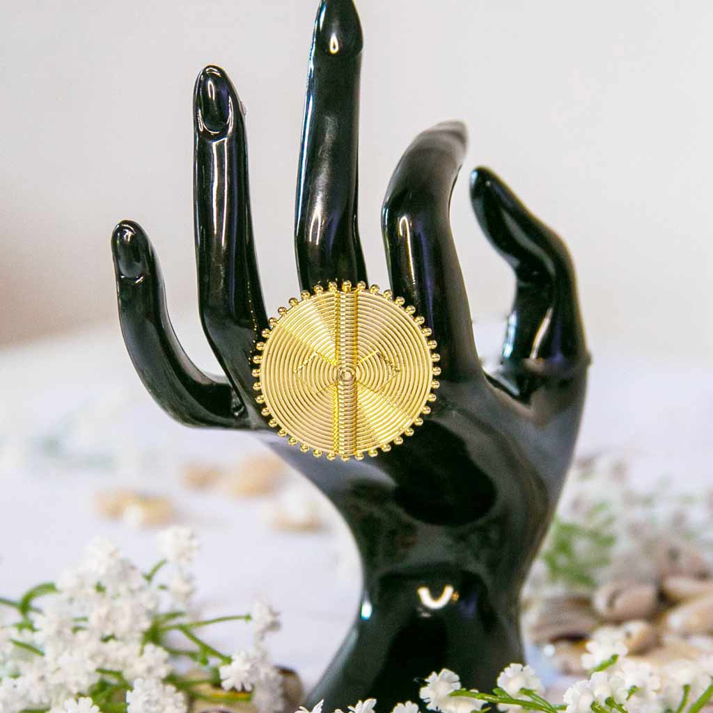 Main de mannequin portant une bague, au majeur,  de style ethnique , ronde et dorée, avec un décor de coquillage cauris et des fleures blanches